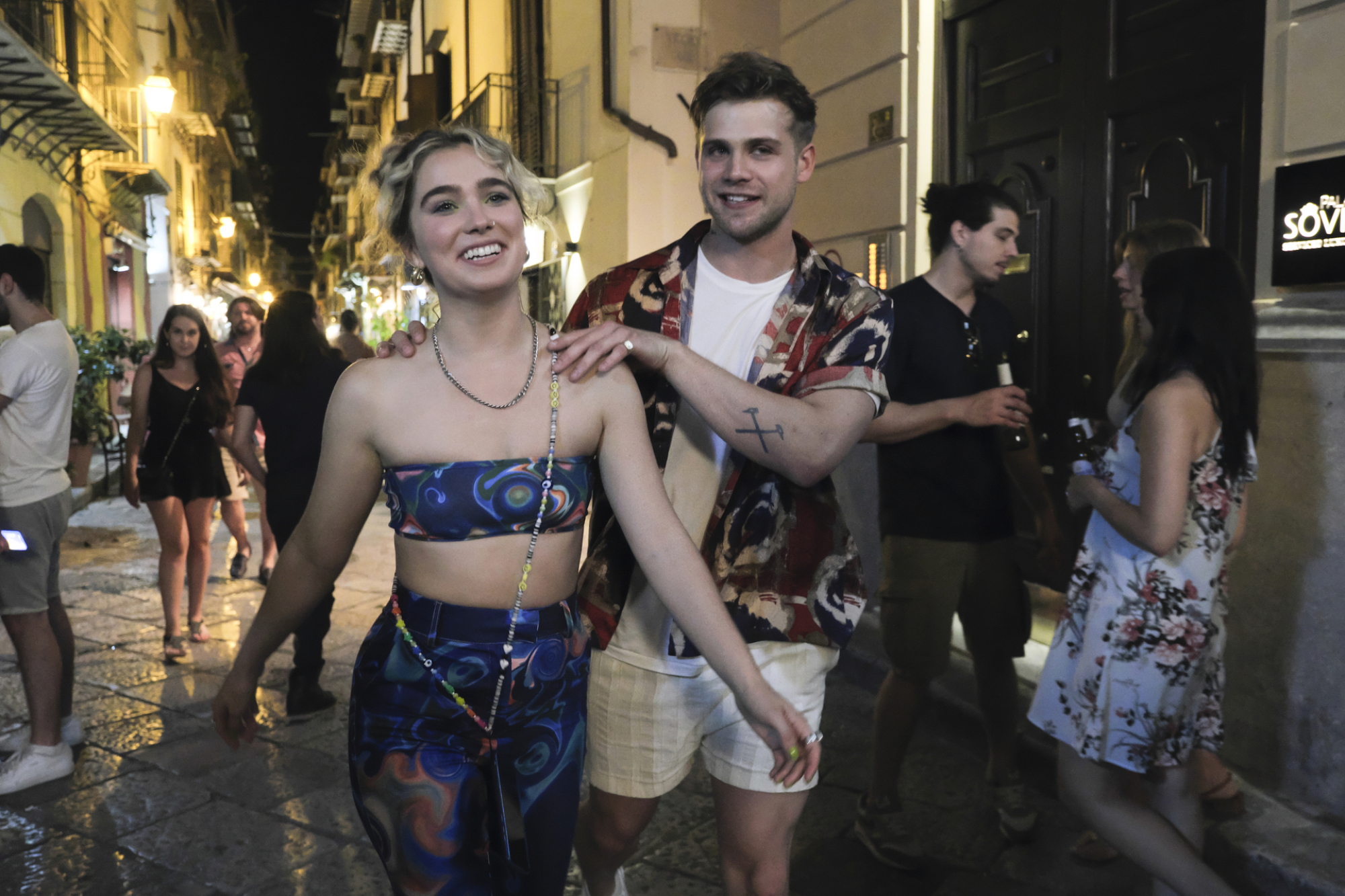 Jack has his hands on Portia's shoulders as they walk down a street in Palermo at night. Portia is wearing a blue matching set. 