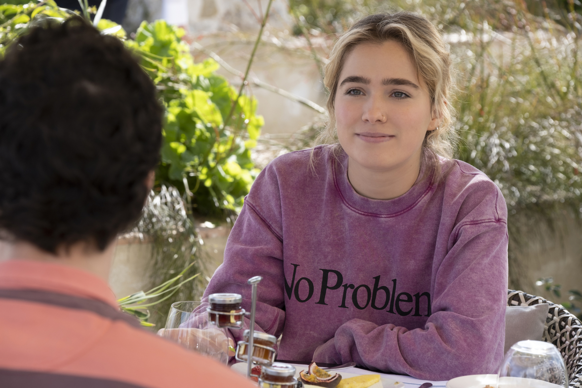 Portia sits at a breakfast table wearing a purple, acid-wash sweatshirt that reads, "No Problemo)