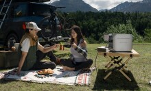 two people sit outside on a picnic blanket in front of mountainous terrain. One holds a cup of coffee and the other holds a pit of coffee. Off to the right is a portable power station, sitting on a folding table