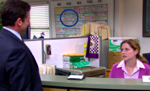 Pam sits at her desk and talks to Michael who is standing at reception.