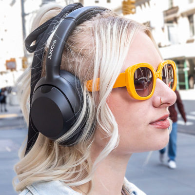 hip young woman in yellow sunglasses wearing Sony headphones on city street