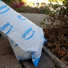 A blue and white Amazon mailer sits on the ground next to a bush. 