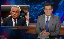 A man in a suit sits behind a talk show desk. In the top-left is an image of another man in a suit.