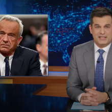 A man in a suit sits behind a talk show desk. In the top-left is an image of another man in a suit.