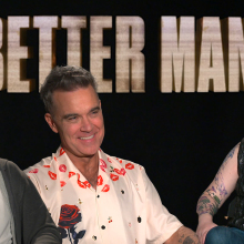 Left to right:  Michael Gracey, Robbie Williams, and Kristy Puchko look at each other, smiling against a black background and 'Better Man' hovering above them