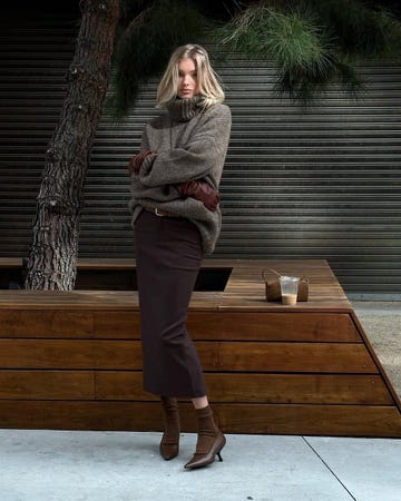 a person stands with arms crossed wearing a chunky sweater a long skirt and heeled boots next to a wooden structure