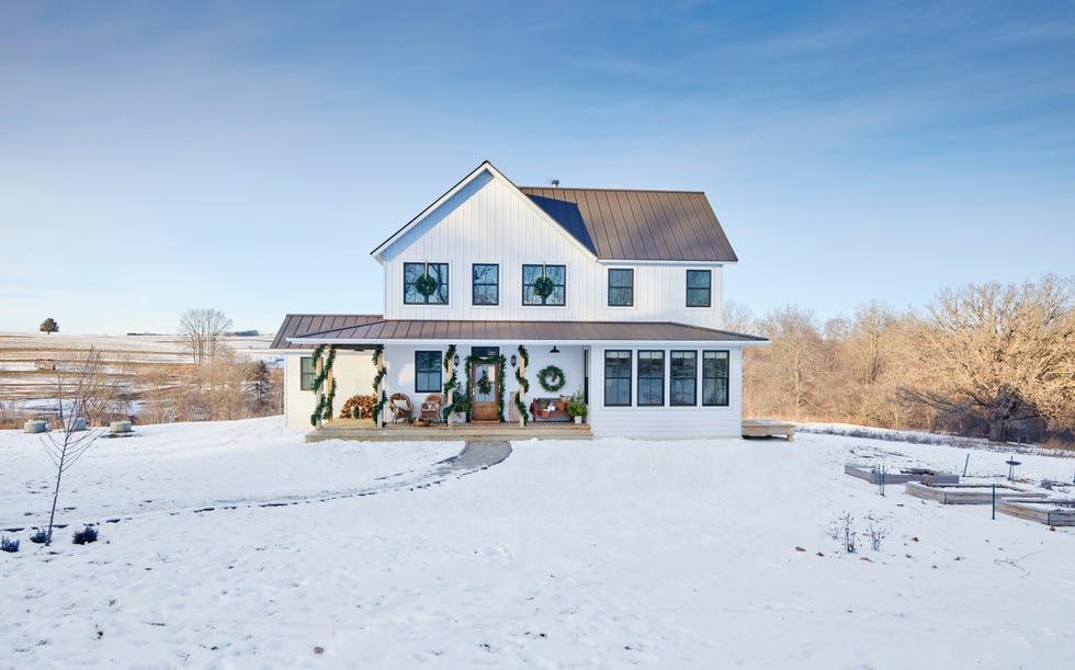 a white farmhouse with christmas greenery on the front porch surrounded by land covered in snow