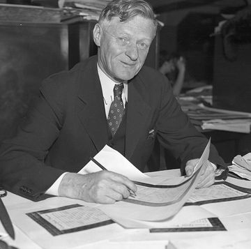 arthur wynne smiles at the camera while seated at a table, he holds a piece of paper and a pencil as several papers rest on the table