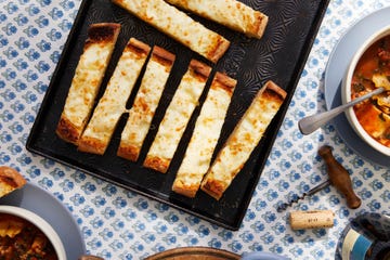 Slices of cheesy garlic bread on a black tray next to a bowl of soup and various utensils