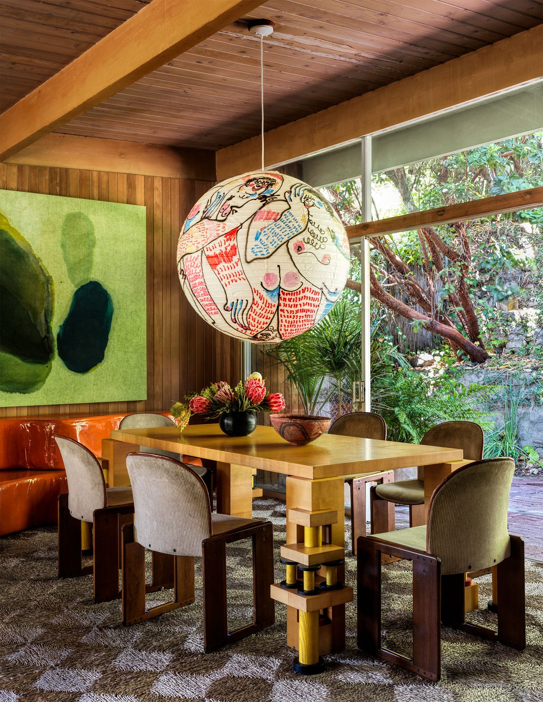 a dining room has wood paneled walls, a chunky wood table with six chairs, a glass wall with door opening to an outside terrace, a large handpainted globe pendant, and a large abstract painting with green hues and shapes