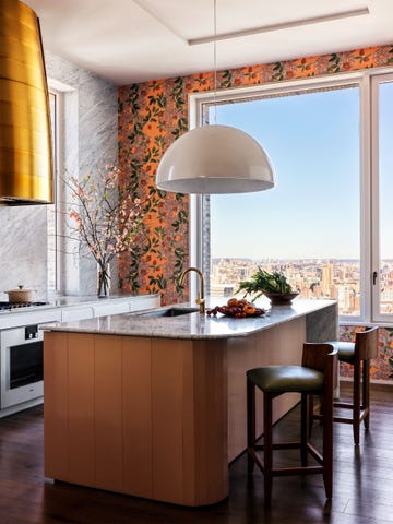 large kitchen window with cityscape view, orange based flowered wallpaper on one wall, marble on wall and countertop, stovetop with brass hood, island with wood base and marble top with sink, large pendant