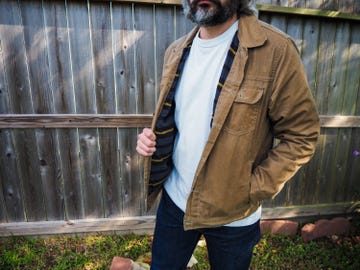 Individual wearing a brown jacket over a white shirt standing against a wooden fence