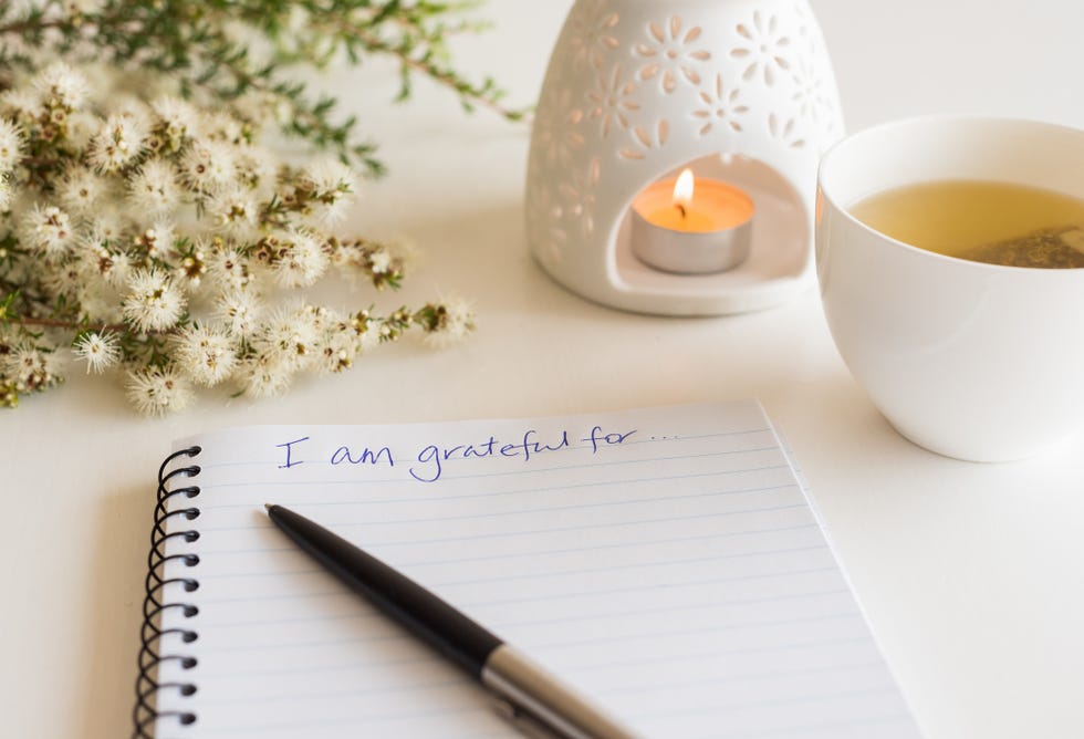 close up of handwritten text i am grateful for in foreground with notebook, pen, cup of tea, flowers and oil burner in soft focus