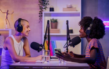 two interracial podcasters are sitting in a small broadcasting studio at home wearing headphones during the discussion and speaking at the microphones two fun girls recording a podcast