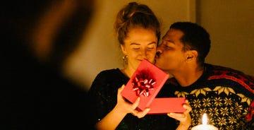 past a shoulder to a couple sitting at a table decorated for christmas with a candle, she opens a red gift box while he kisses her on the cheek