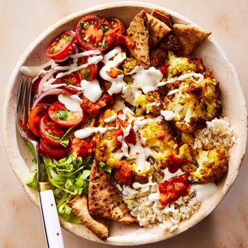 roasted cauliflower in a bowl with tomato salad, crushed pita, onions on top of rice and drizzled with white halal cart style sauce