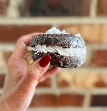 a hand holding a chocolate donut with white cream filling set against a brick background