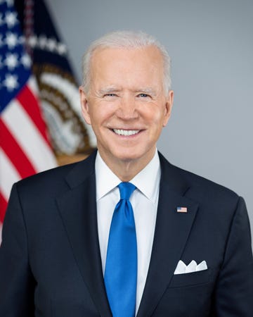 joe biden smiles at the camera while standing in front of a blurry american flag, he is wearing a navy suit, a white collared shirt, a royal blue tie, and an american flag pin on his lapel, he has a white pocket square in his breast pocket