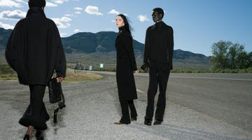 three figures in black clothing standing on a roadside with mountains in the background