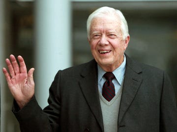 jimmy carter smiles and waves to the camera, he is wearing a blue and white striped collared shirt with a navy and dark red diagonal striped tie, a light gray v neck sweater is on top of the shirt and tie, and he also wears a dark gray suit jacket