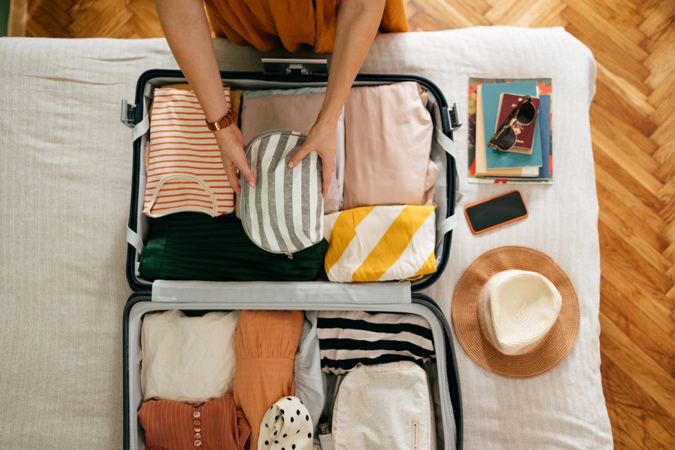 shot from above of an anonymous woman packing things in her suitcase on the bed she is holding and putting a grey and white stripped cosmetics bag in there is her mobile phone, books and sunglasses on the bed