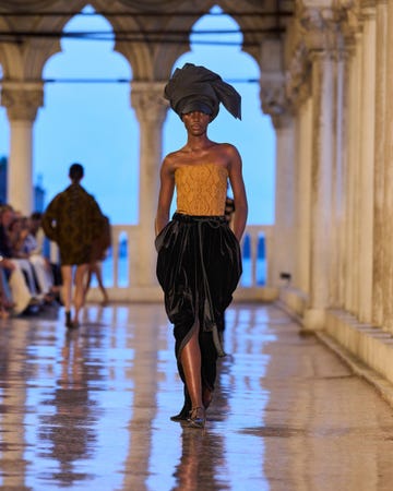 a model walks down a runway wearing a strapless top in a lace pattern, paired with a long, flowing skirt made from black velvet the models hair is styled with a large headwrap the runway is set in a grand architectural space with arched windows, reflecting soft natural light, creating a serene atmosphere additional figures in stylish attire can be seen in the background, observing the performance