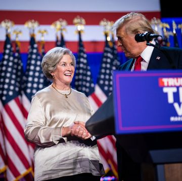 susie wiles smiles at donald trump as they shake hands on a stage, many american flags stand in the background, a podium with a microphone is in the right side of the frame