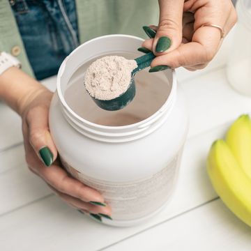 woman in jeans and shirt holding measuring spoon with portion whey protein powder above plastic jar on white wooden table with shaker, banana and apple fruit process of making protein drink