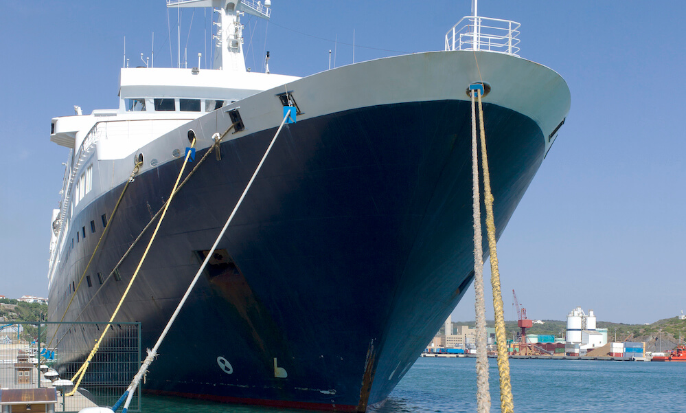 A large boat moored in the harbor as seen from below. It is a sunny day and the sea is clear and calm.In the foreground, the ropes of the boat