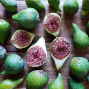 Many green figs are laid out on a wooden surface, a few of the figs have been cut down their middle revealing their bright red flesh.