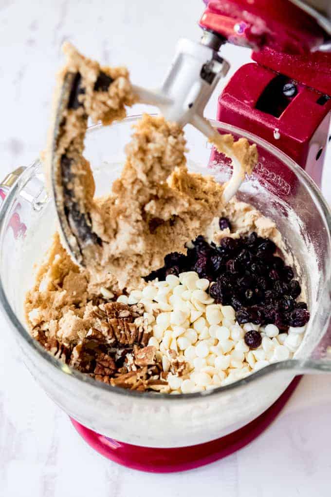 Cookie dough in the bowl of a mixer with white chocolate chips, pecans, and dried cranberries.