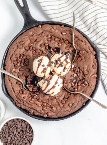 An overhead image of a chocolate skillet cookie with three spoons in it and ice cream on top.