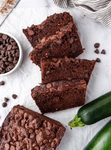 Top view of slices of chocolate zucchini bread on a white surface with zucchini and chocolate chips around them.