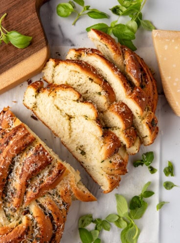 An overhead image of a loaf of sliced bread surrounded by fresh basil leaves, parmesan cheese, and a wooden cutting board.