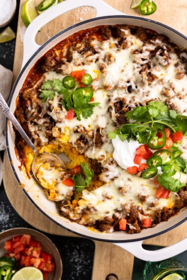 White baking dish with tamale pie topped with melted cheese, sliced jalapeno, diced tomatoes, sour cream and cilantro.