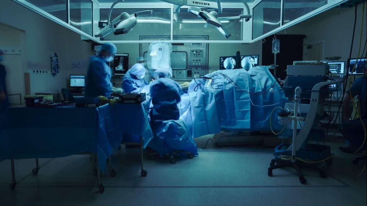 An operating room, with patient - covered in blue surgical drapes - in the centre, with two seated surgeons working on their ankle. In the background there are several screens showing X-Rays on an ankle. In the foreground there is a table covered with a blue drape.