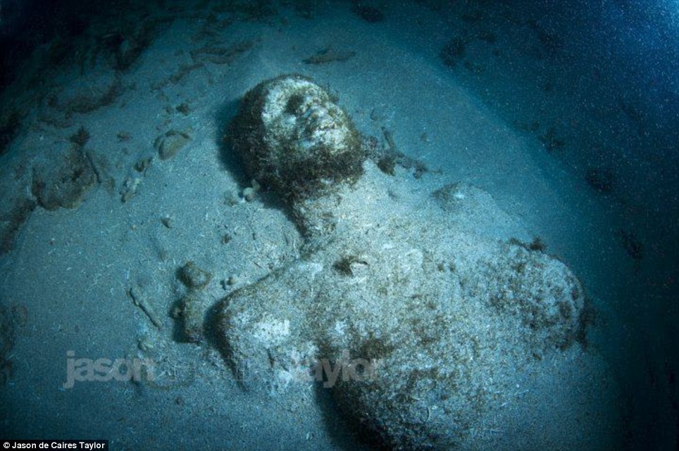 From above: The head and torso of a woman is seen on her back as she is gradually covered by plants and debris that still fail to hide her distinct curves and features