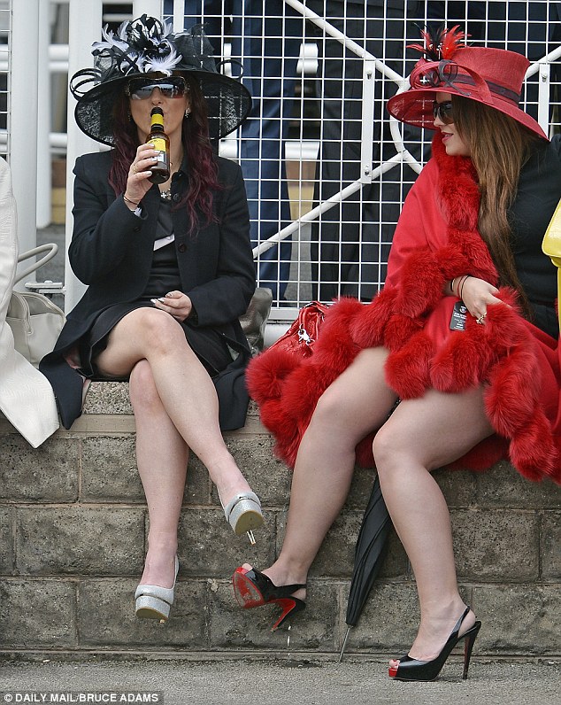 Two Ladies' Day visitors to the racecourse, both in big hats and heels, one wearing a bright red fur cape while another wears black