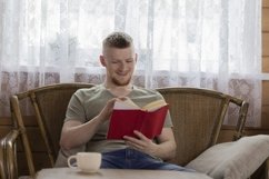 young man reading book with red cover on wicker bench Product Image 1