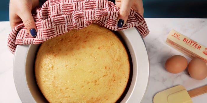 Hands holding a KitchenAid cake pan with a baked cake inside next to a yellow spatula, two eggs, and a stick of butter.