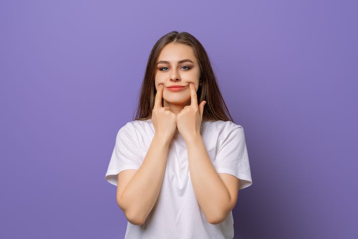 A stock image of a woman holding up sides of her mouth to make a fake smile