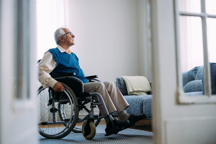 An elderly man sits thoughtfully in a wheelchair in a bright living room. He gazes out, possibly reflecting on past memories. The scene is serene and contemplative.