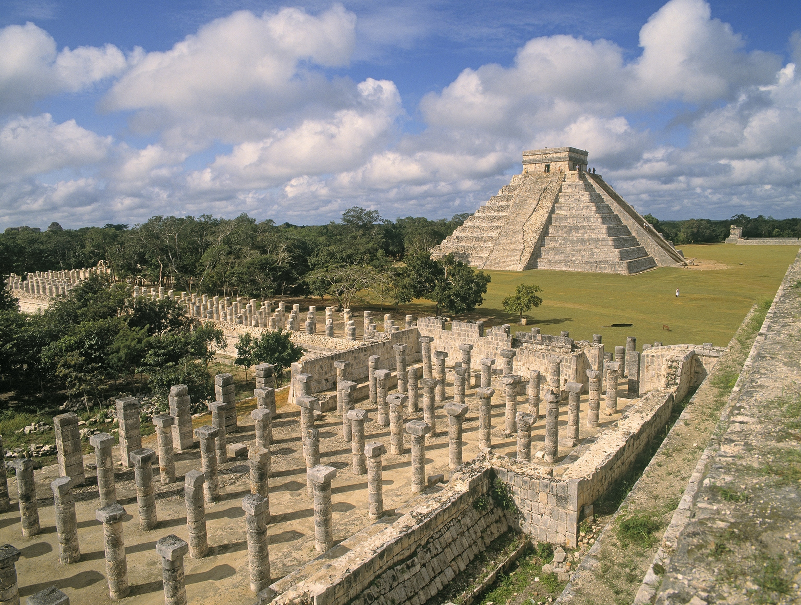 Cloudy blue skies hover over the stone pyramid, with an abundance of columns in the foreground, and green grass surrounding the structure.