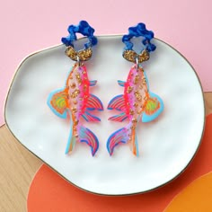 Laser cut fish earrings featuring a unique hand painted pattern of orange, pink, yellow, purple, blues and gold glitter resin with tiny hand painted details. The fish dangle from a small blue and gold glitter top piece.- Painted pattern is over clear, see through, acrylic. This effect gives the earrings a fun layered depth.- Earrings are 2.75 inches long by 1.25 inches wide. - Earrings are push back studs in hypoallergenic stainless steel. - Lightweight, not heavy at all. Great for daily wear.- Hand Painted Pattern, Fish Earrings, Glitter Top, Mixed Media Jewelry, Orange And Gold, Clay Earring, Plastic Crafts, Dope Jewelry, Jewelry Fashion Trends