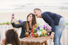 a man and woman taking pictures with their cell phones at a table on the beach