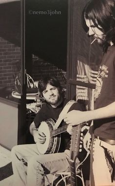 a man sitting down playing a guitar with another man standing next to him and looking at the camera