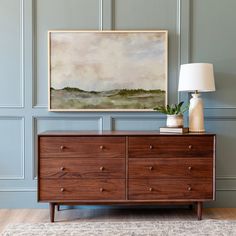 a painting hangs on the wall above a dresser with a lamp and vase in front of it