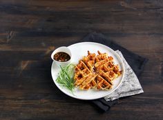 a white plate topped with waffles next to a bowl of sauce on top of a wooden table