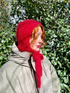 a woman wearing a red knitted hat and scarf in front of some green bushes