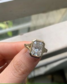 a woman's hand holding an engagement ring with a diamond in the middle and a window behind it